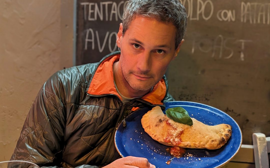 Man in a jacket holds a blue plate with a calzone and basil leaf.