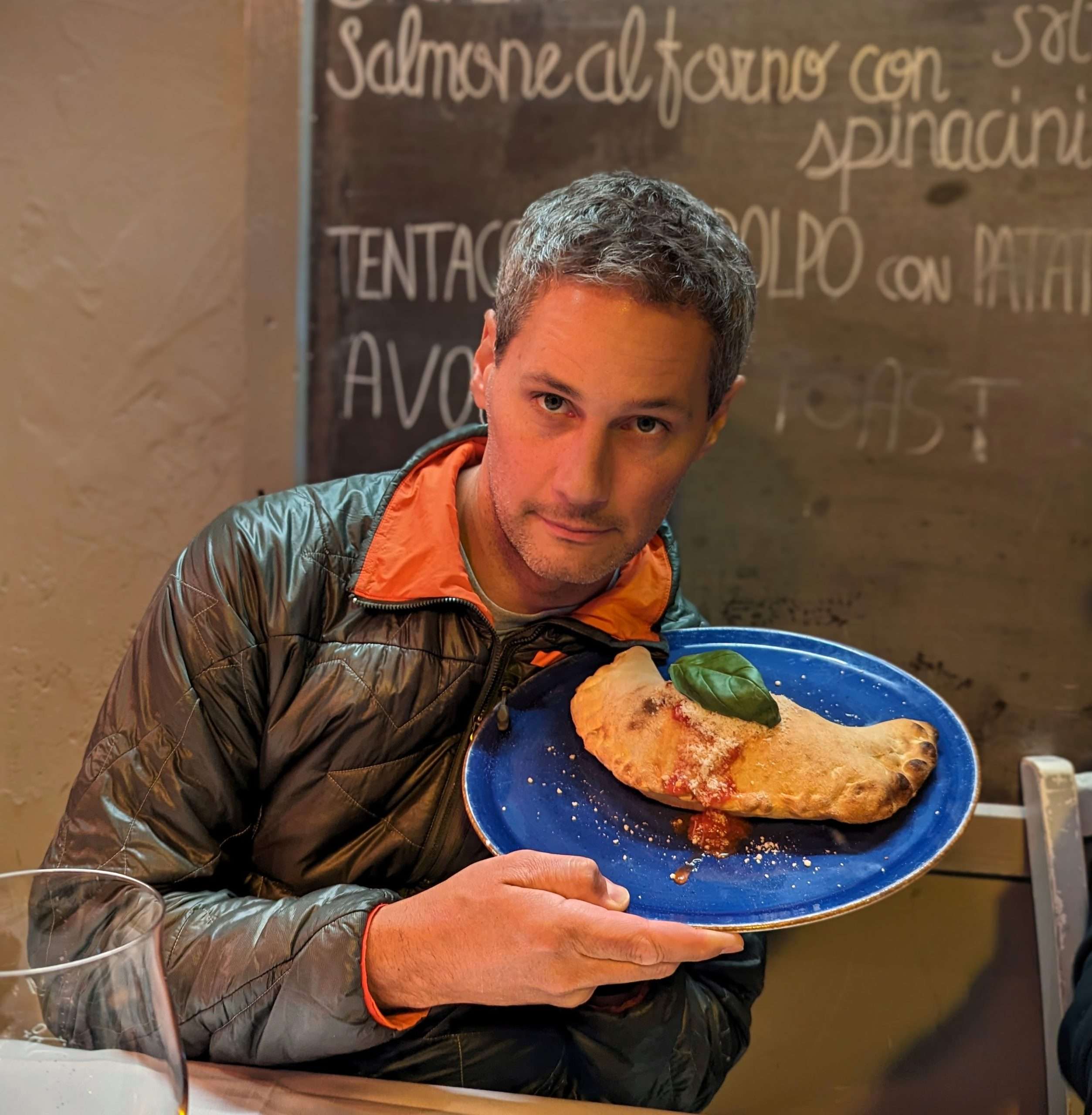 Man in a jacket holds a blue plate with a calzone and basil leaf.
