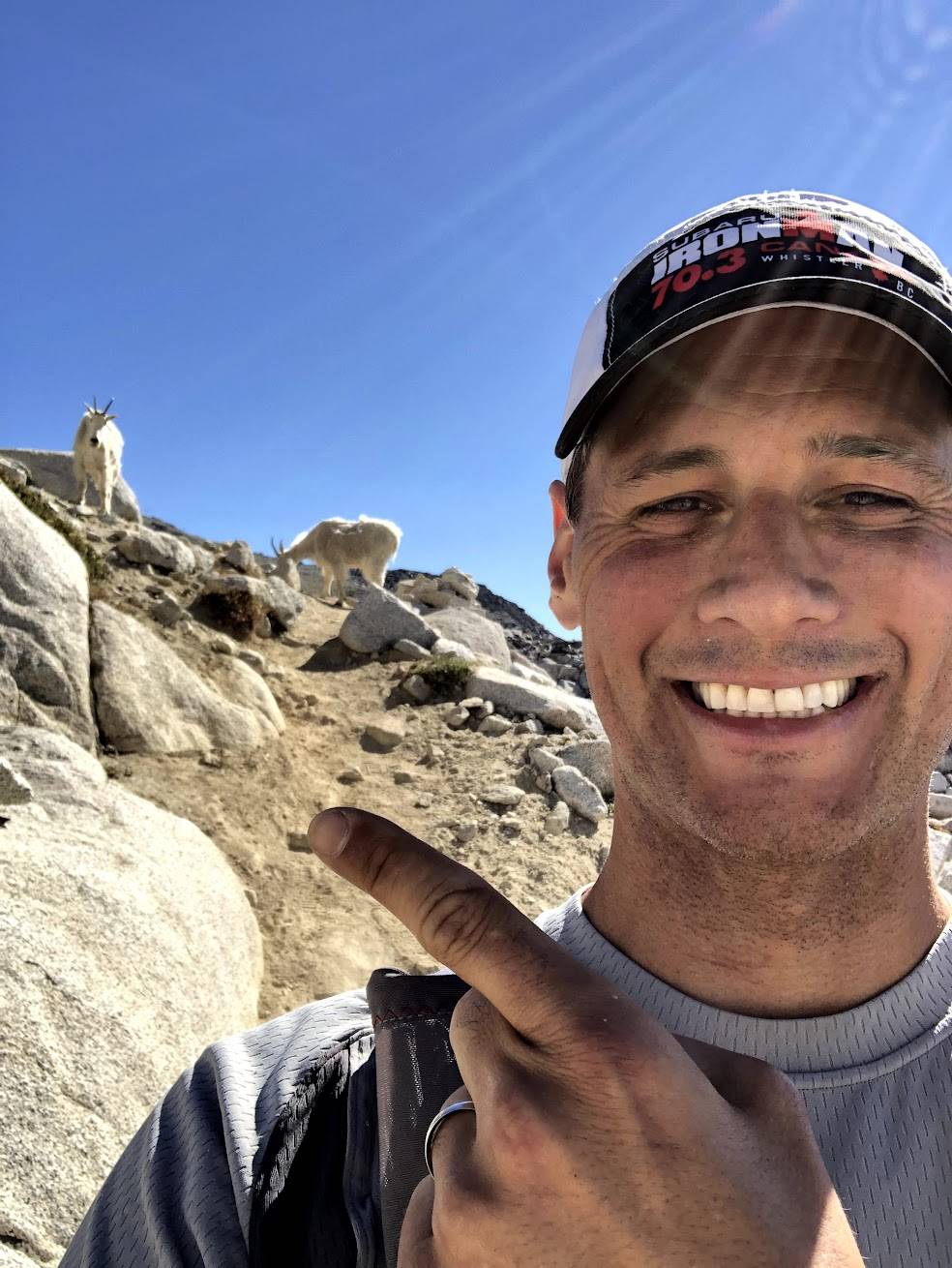 Smiling man on rocky mountain with goats in background under blue sky.