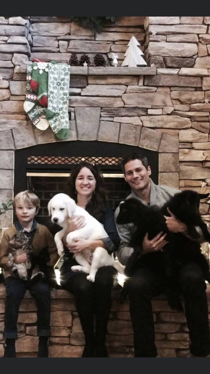 Family with two dogs sits on a stone fireplace mantel, smiling for the photo.